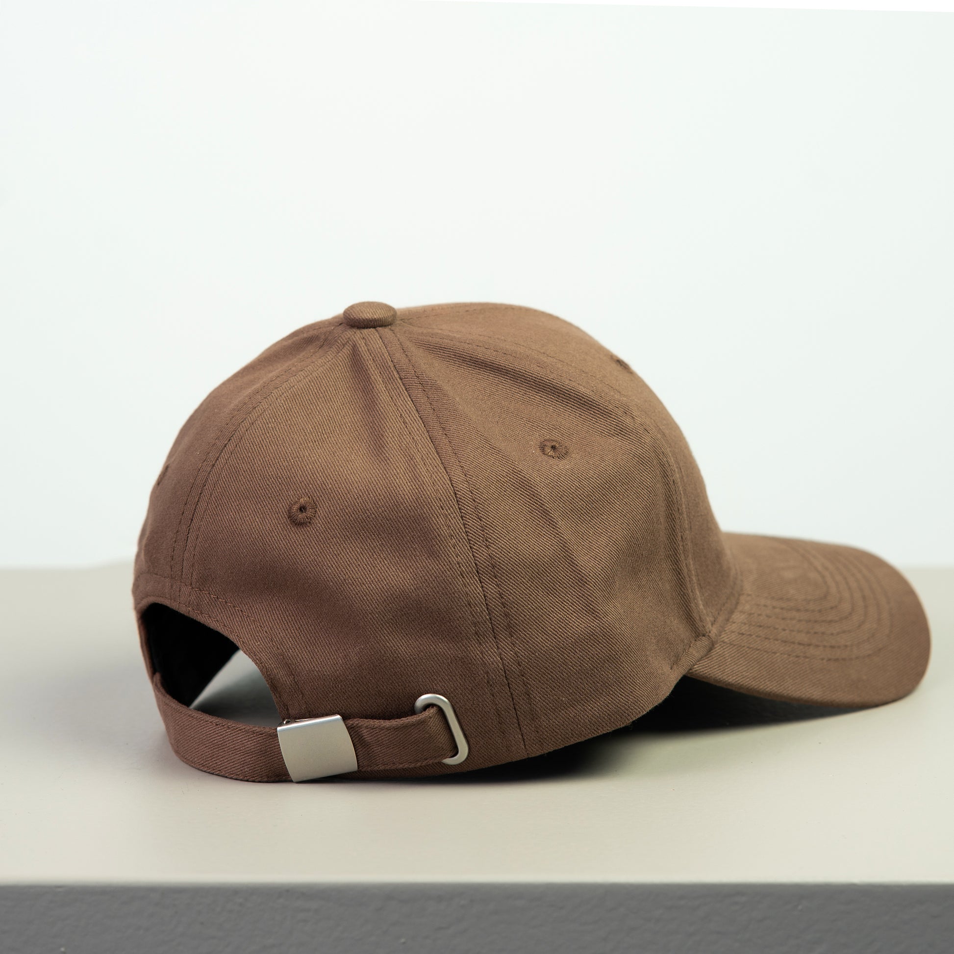 Brown baseball cap with adjustable metal buckle on a light gray surface with a white background