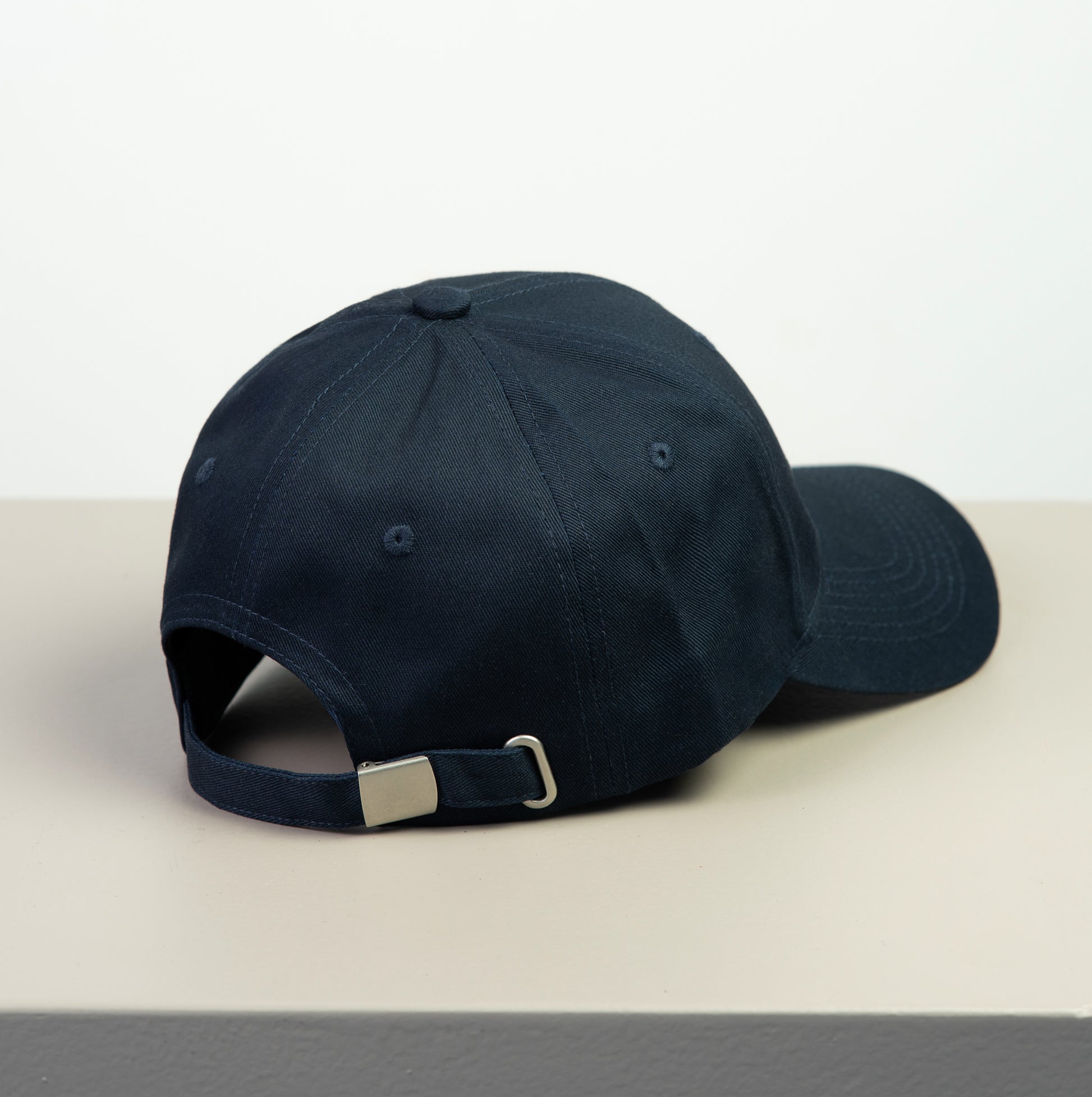 Navy blue baseball cap with metal adjustment buckle on a light gray surface with a white background
