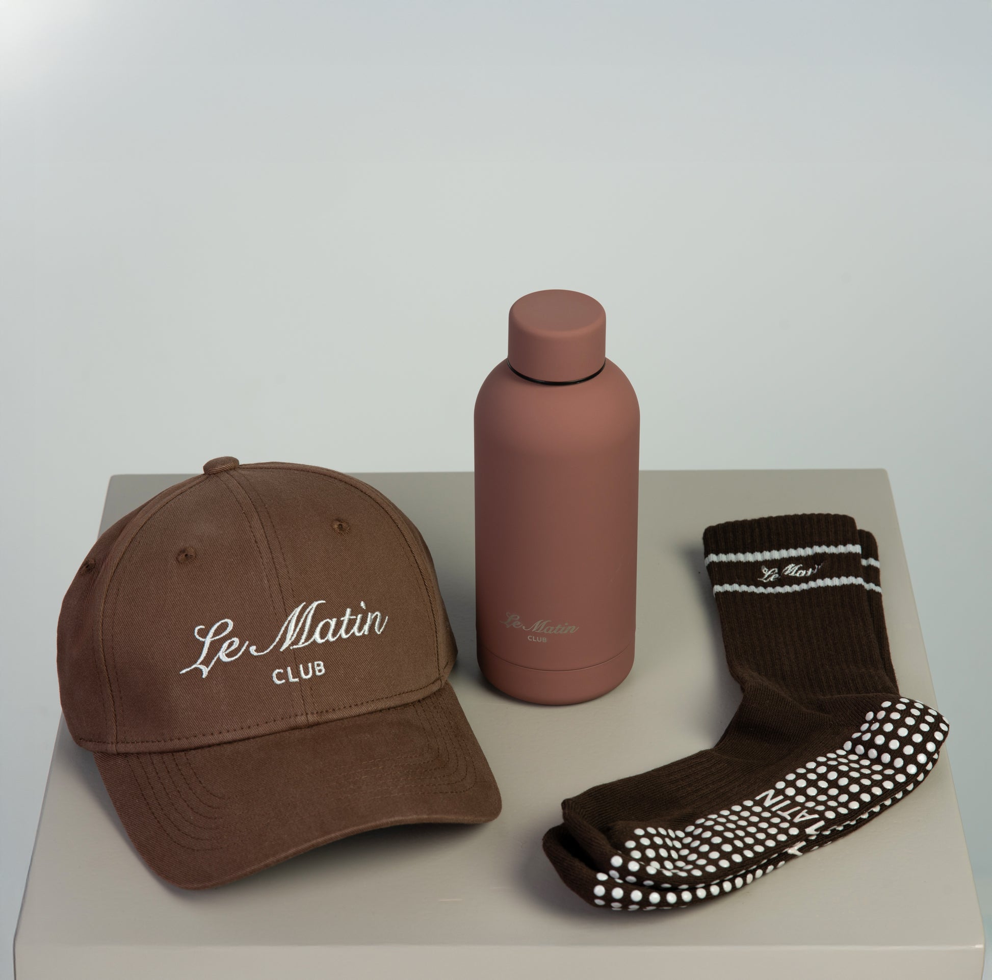 Brown cap, brown water bottle, and brown socks with grey on a light gray surface with Le Matin branding.