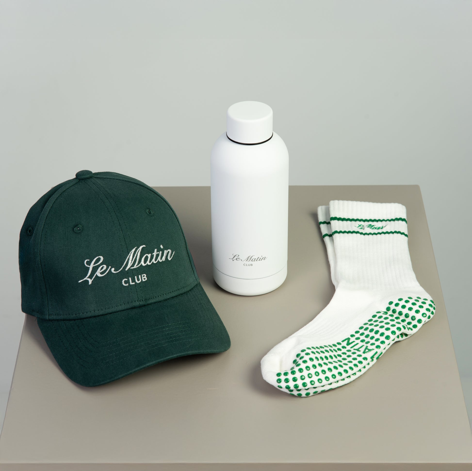 Green cap, white water bottle, and green and white socks on a gray surface with 'Le Matin Club' branding.